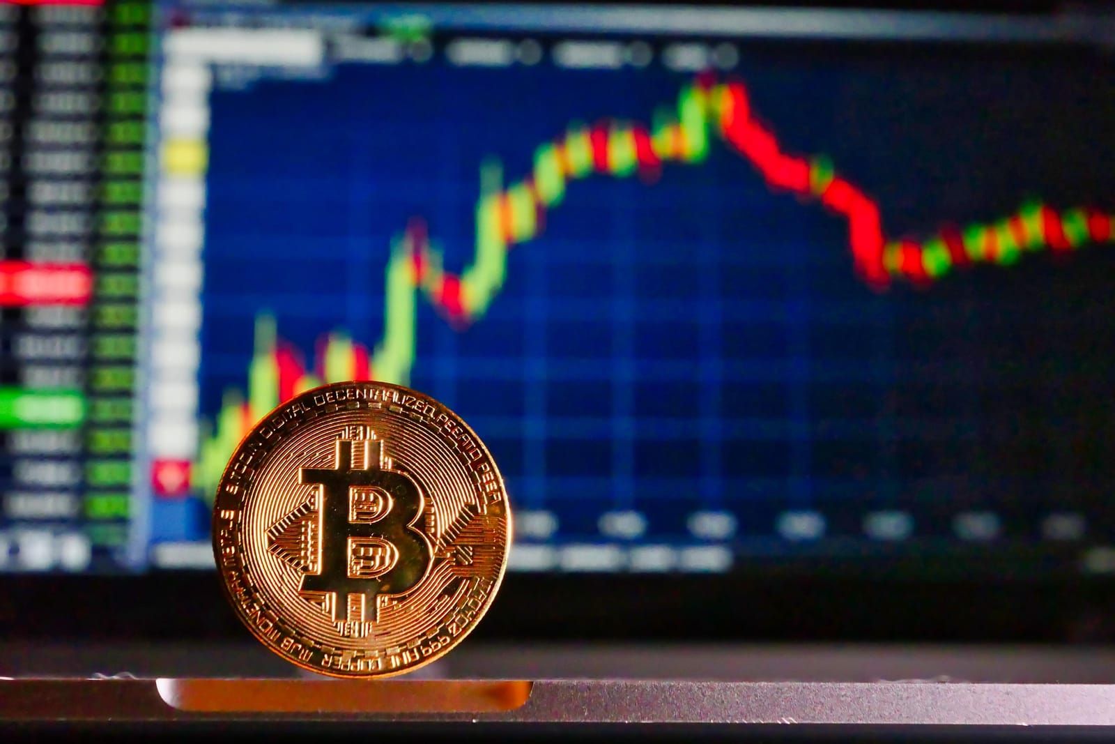 A single bitcoin coin in front of a blurred screen showing a red and green candlestick chart